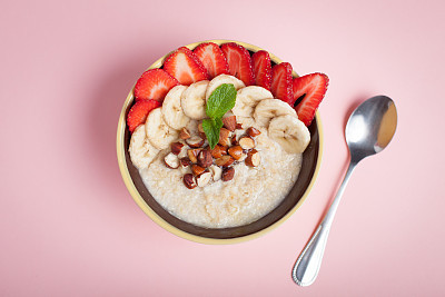 Bowl of oatmeal with a banana, strawberries, almonds, hazelnuts and butter on a pink background. Hot and a healthy dish for Breakfast, top view