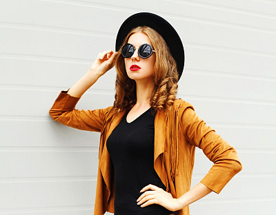 Fashion woman in black round hat, sunglasses, jacket on gray wall background