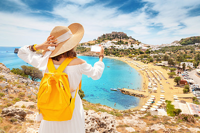 Young and happy woman tourist taking selfie photo on her smartphone with view on a sandy beach and old town. Vacaction and travel concept