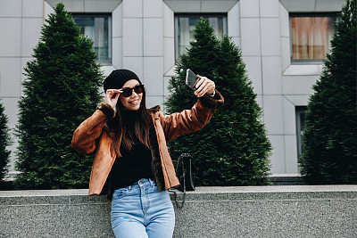 City style. Technology. Autumn/spring. Stylish girl in casual clothes is doing selfie using a smartphone and smiling while walking outdoors