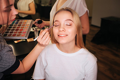 Nice young woman sit in beauty room with closed eyes. She smile. Make up artist put eyeshadows on eyeline. She hold palette in hands. Another profesional stand behind.
