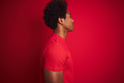 Young american man with afro hair wearing t-shirt standing over isolated red background looking to side, relax profile pose with natural face with confident smile.