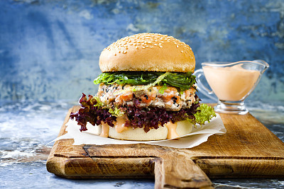 Asian style salmon burger with grilled shrimps, seaweed, lettuce and spicy sriracha mayo sauce served on pieces of brown paper on a rustic wooden board. Bright blue background with copyspace