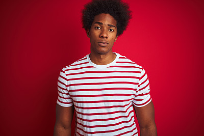 Young african american man with afro hair wearing striped t-shirt over isolated red background Relaxed with serious expression on face. Simple and natural looking at the camera.