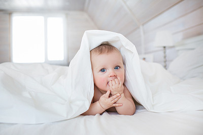 Pretty Infant lies under a white cover in bed in bedroom