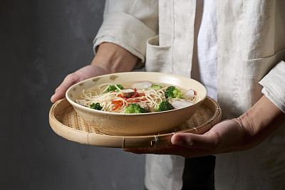 Two hands holding a bowl with vegan wok noodles with broccoli, cherry tomato, garlic, onion, black olives and olive oil flavored with salt, peppers, chili, turmeric and soy sauce