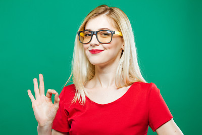 Cute Smart Girl in Eyeglasses is Showing Ok Sign on Green Background. Beautiful Blonde with Long Hair and Red Top in Studio.
