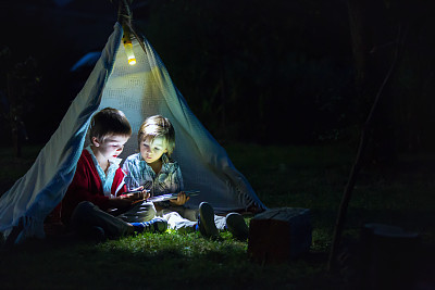 Cute little brothers, playing on tablet and telephone at night in campside, in the tent