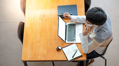 Young Asian man university student using smartphone while working with laptop computer and lecture notebook on wooden table in the college. Campus lifestyle in education building. Shot from top view