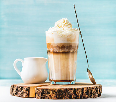 Latte macchiato with whipped cream, serving silver spoon and pitcher on wooden round board over blue painted wall background
