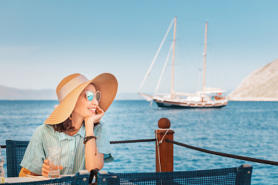 Happy woman wearing big hat sitting in cafe and looking to the seascape