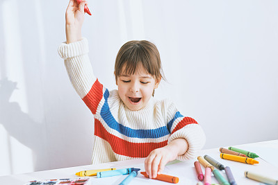 Happy little girl smiling and playing with colorful pencils. Joyful child drawing with colorful pencils on paper at the desk, feeling happy, sitting at home. People, childhood, education