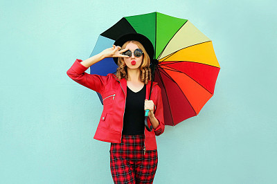 Stylish young woman with colorful umbrella blowing red lips sending sweet air kiss, wearing red jacket, black hat on blue wall background