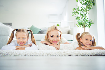 Portrait of three nice cute lovely adorable attractive charming cheerful cheery people pre-teen girls mom mum mommy lying on carpet in light white interior room house indoors