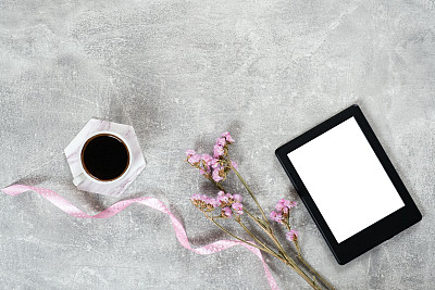 Flat lay elegant feminine accessories: coffee cup, pink ribbon, dry flowers and e book reader on concrete surface. Top view beauty blogger workspace, freelancer home office desk. Fashion blog hero.