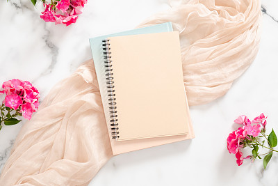 Composition with blank paper notebook, pink flowers and pastel blanket on marble background. Flat lay, top view still life concept.