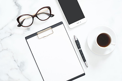 Modern home office desk workspace with blank paper clipboard, glasses, stationery, smartphone on white marble background. Flatlay, top view.