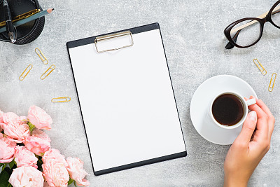 Stylish woman desk with blank paper clipboard mockup, pink rose flowers, gift box with gold color ribbon, coffee cup, glasses on stone background. Home office desk, beauty blogger workspace concept