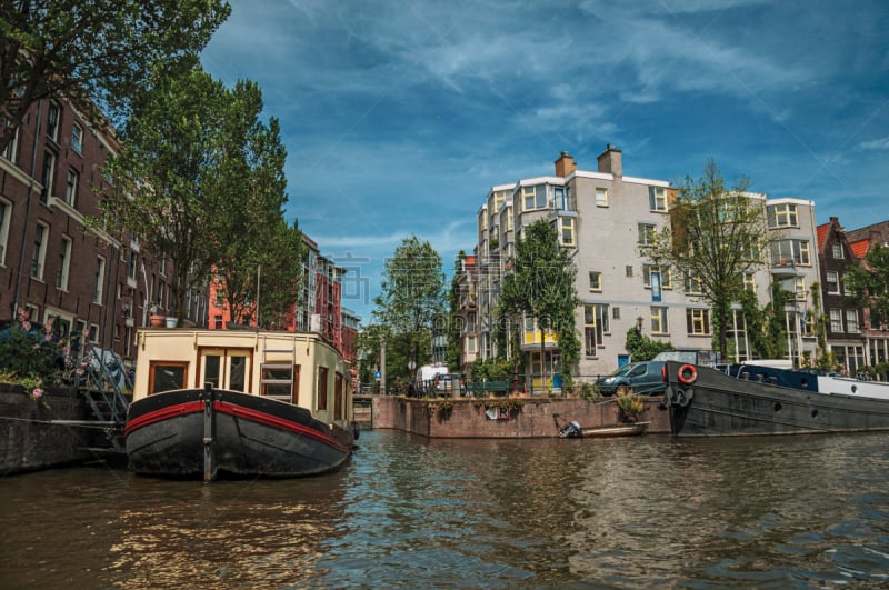 Big boat used as house moored at side of tree-lined canal with old buildings and sunny blue sky in Amsterdam.