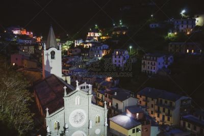 RIOMAGGIORE ITALY, Light catches façade and tower of church on hill at night with colors of street and homes light in background