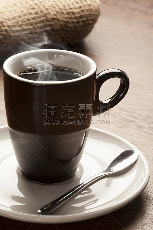 Xícara preta com café quente, em pires com colher, sobre mesa de madeira