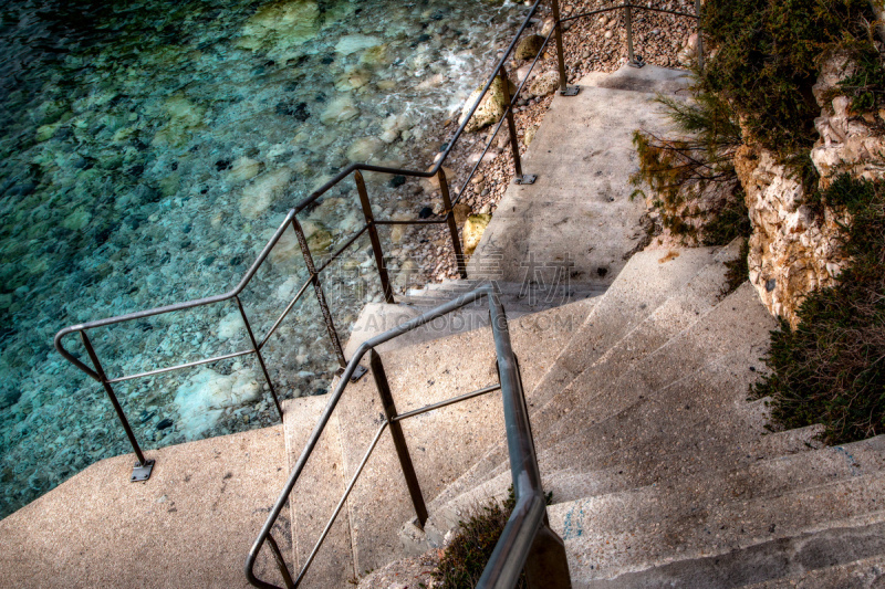 Un escalier d'accès à la mer sur le littoral marseillais.
Des marches en béton mènent vers une eau très transparente. 
L'image s'ouvre sur la rampe métallique que l'on suit jusqu'en bas.
L'image est fermée par les rochers de la côte.
Photo en couleur