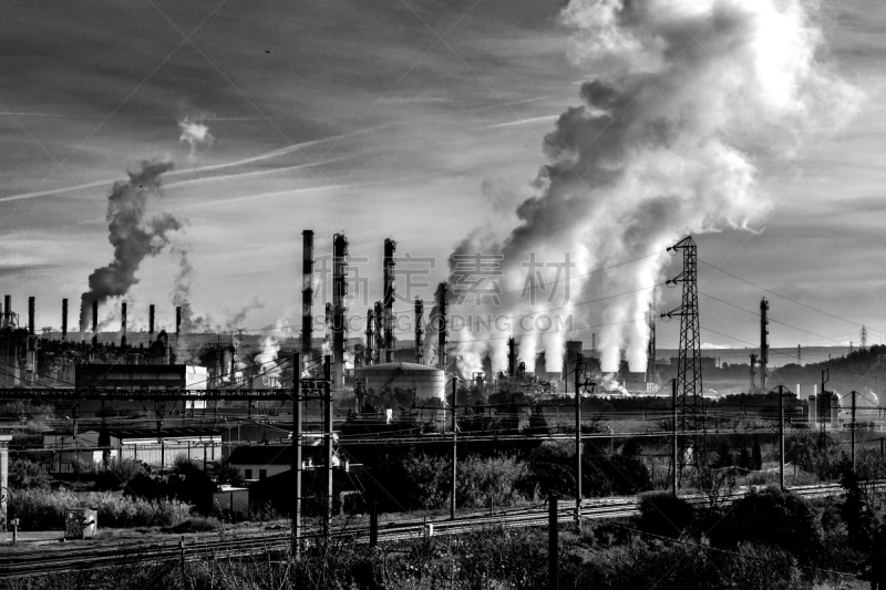 Vue extérieure de l'usine pétrochimique de Berre (sud de la France).
En premier plan, une voie ferrée qui longe l'usine.
D'énormes nuages de fumée sortent des cheminées
Sur la gauche, l'image s'ouvre sur une série de cheminées.
Image en noir et blanc