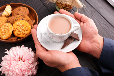 A man is holding a cup of coffee. Nearby are cookies and a flower. Indoors.