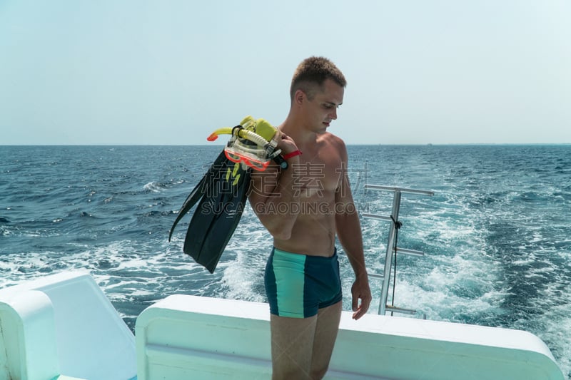 The guy stands in the slats in the stern of the yacht with fins and a mask in his hands. The guy is preparing to dive under water