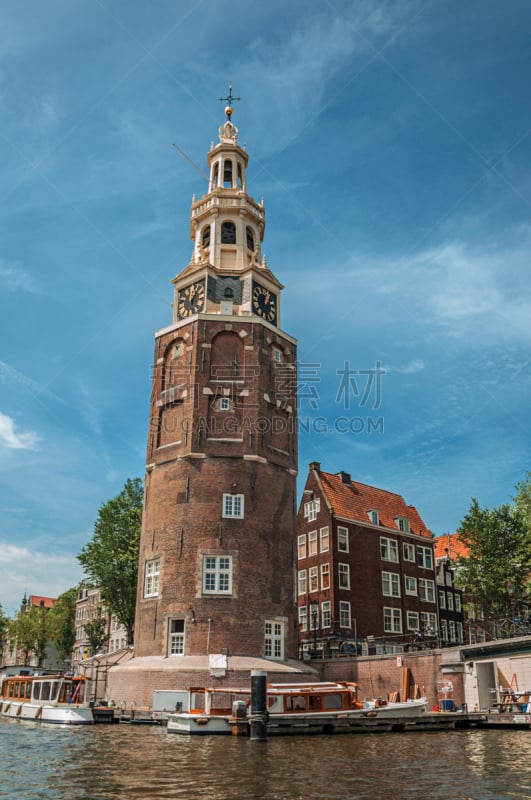 Old brick bell tower and buildings near the tree-lined canal with moored boats and blue sky in Amsterdam.