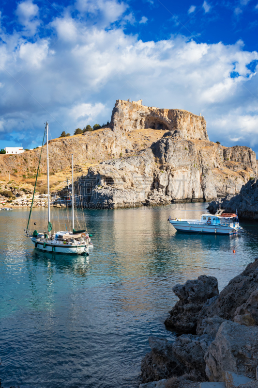 Sail boats in St. PaulÂ´s bay, cloudy blue sky, Lindos acropolis in background (Rhodes, Greece)