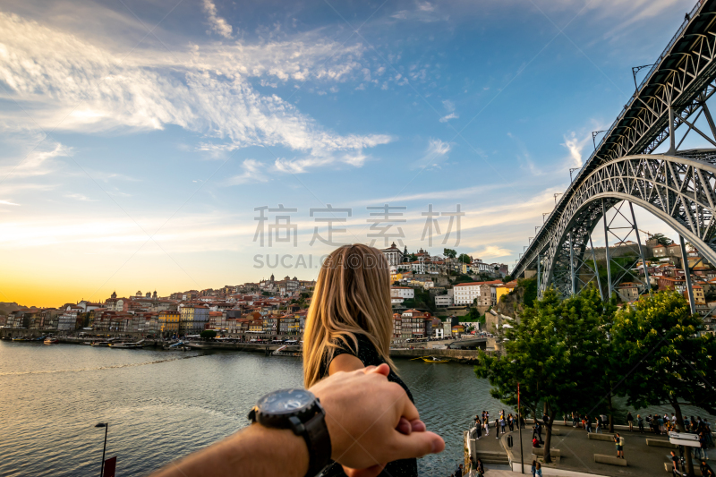 A woman tends back her hand into the hand of her husband. In the background, blue skies and soft white clouds roam over the Douro river between the cities of Porto and Vila Nova de Gaia displaying it's colourful City-line. The double decked metal arched b