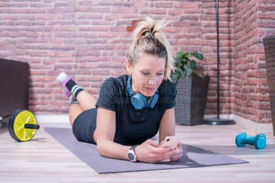 Girl, with blonde hair, does sports at home in the salon. Only because of the insulation for COVID19. She wears sports clothes and wireless headphones, is lying on a mat and smiling while chatting on the phone.