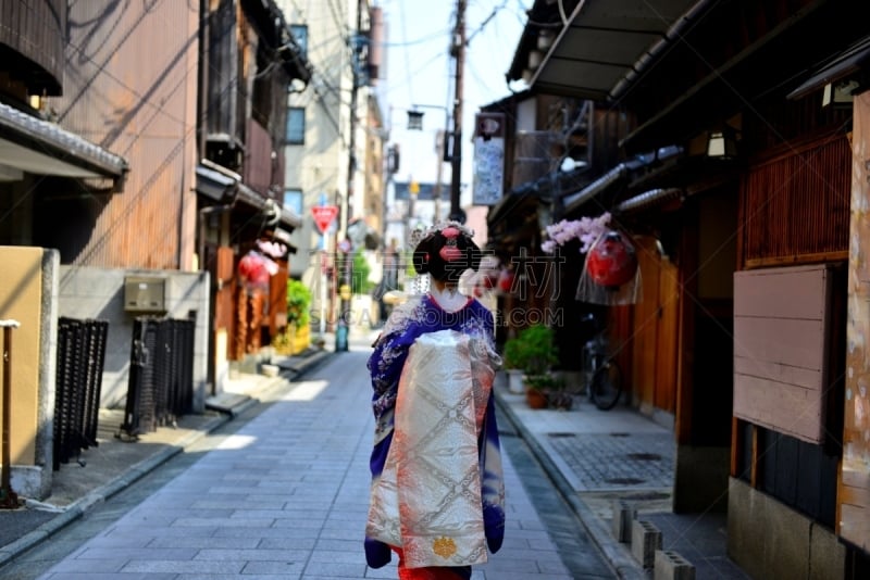 A Japanese woman in Maiko’s costume and hairstyle is walking on the street of Miyakawa-cho, Gion, Kyoto. She wears traditional long-sleeved kimono with long dangling sash and her hair is elaborately decorated with seasonal flower-featured hairpins.