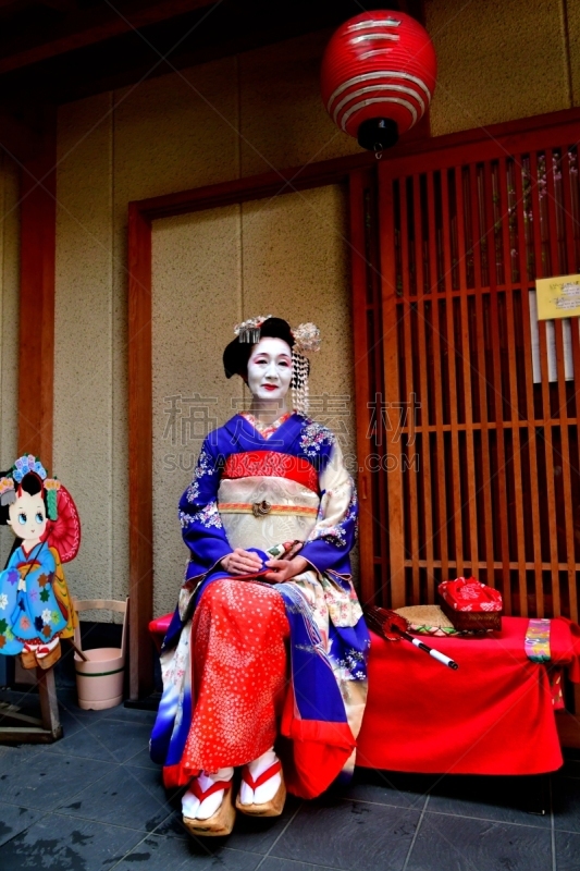 A Japanese woman in Maiko’s costume and hairstyle is resting on the bench in the courtyard of Geisha House in Miyakawa-cho, Gion, Kyoto. She wears traditional long-sleeved kimono with long dangling sash and her hair is elaborately decorated with seasonal 