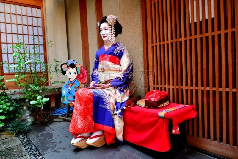 A Japanese woman in Maiko’s costume and hairstyle is resting on the bench in the courtyard of Geisha House in Miyakawa-cho, Gion, Kyoto. She wears traditional long-sleeved kimono with long dangling sash and her hair is elaborately decorated with seasonal 