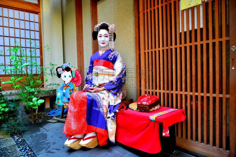 A Japanese woman in Maiko’s costume and hairstyle is resting on the bench in the courtyard of Geisha House in Miyakawa-cho, Gion, Kyoto. She wears traditional long-sleeved kimono with long dangling sash and her hair is elaborately decorated with seasonal 