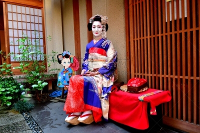 A Japanese woman in Maiko’s costume and hairstyle is resting on the bench in the courtyard of Geisha House in Miyakawa-cho, Gion, Kyoto. She wears traditional long-sleeved kimono with long dangling sash and her hair is elaborately decorated with seasonal 